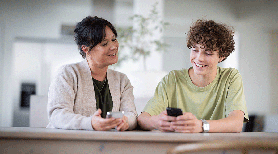 mor og sønn sitter ved bord og smiler, de ser på mobiltelefonen til sønnen