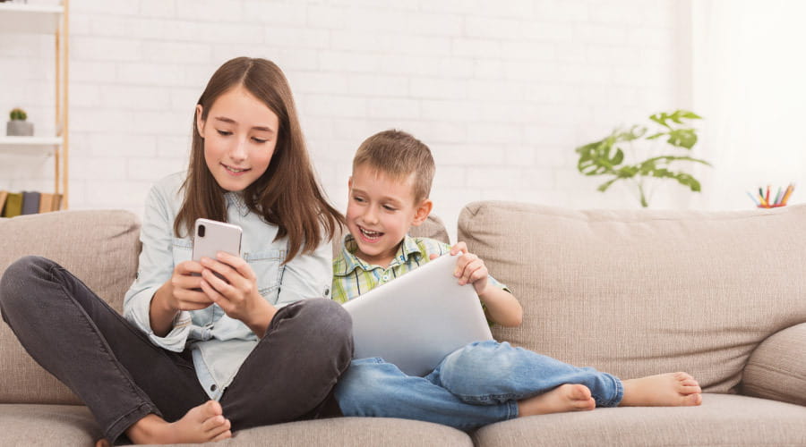 To barn som sitter i sofa med nettbrett og telefon