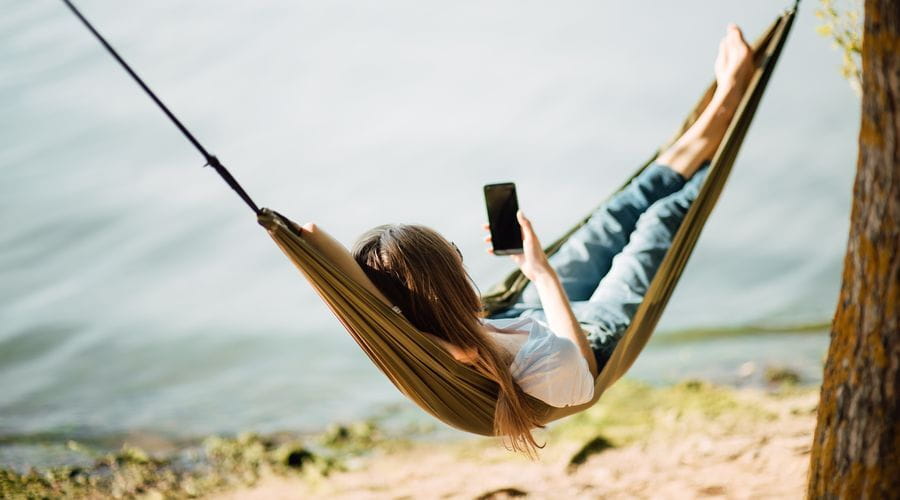 Jente ligger i en hengekøye ved et vann og ser på mobilen sin