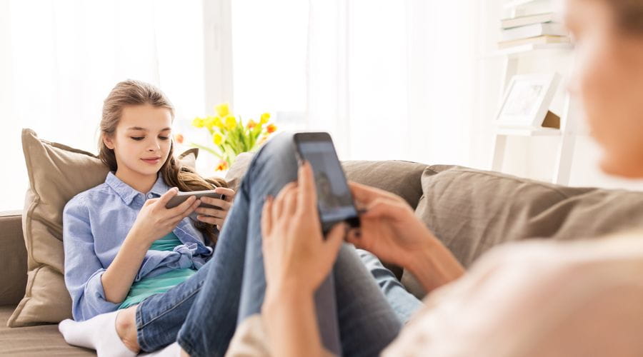 Jente som sitter i sofa med en telefon, mens mor sitter på andre siden med sin egen telefon