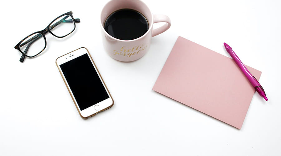 En telefon, ett par briller, en kopp kaffe og en rosa notatbok som ligger på et bord