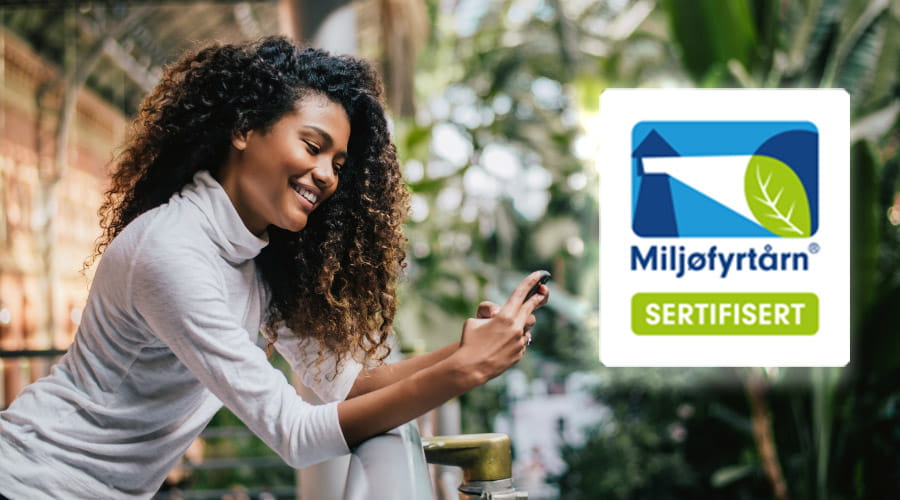 Dame som står i et drivhus fylt med planter som ser på mobilen sin. Redigert inn Miljøfyrtårnsertifisering